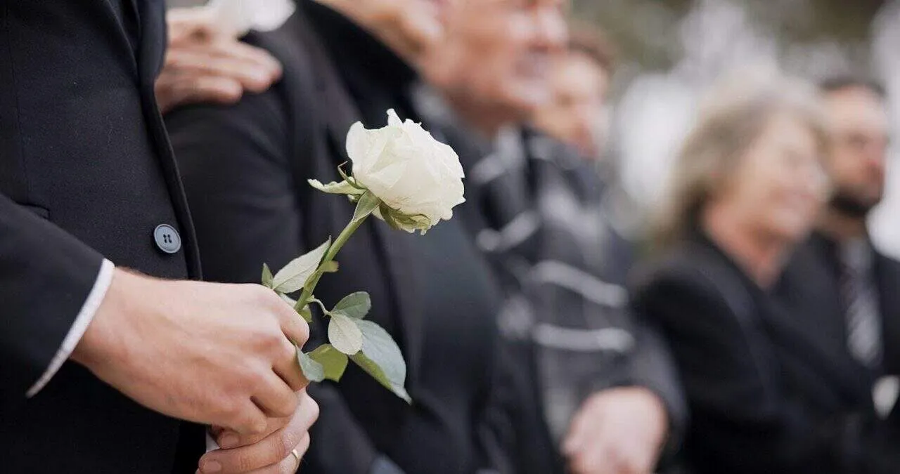 Close-up of hand holding white rose during memorial service with loved ones gathered in remembrance