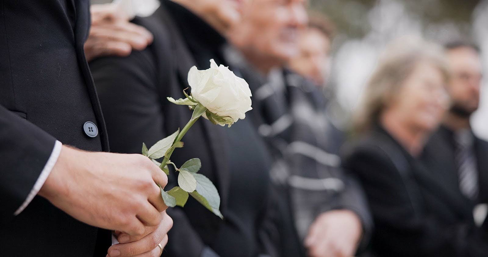 Close-up of hand holding white rose during memorial service with loved ones gathered in remembrance
